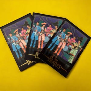 May include: Three black and gold trading cards featuring a photo of the Beatles performing on stage. The cards have a psychedelic background with swirling colors and the text "The Beatles" in gold lettering.