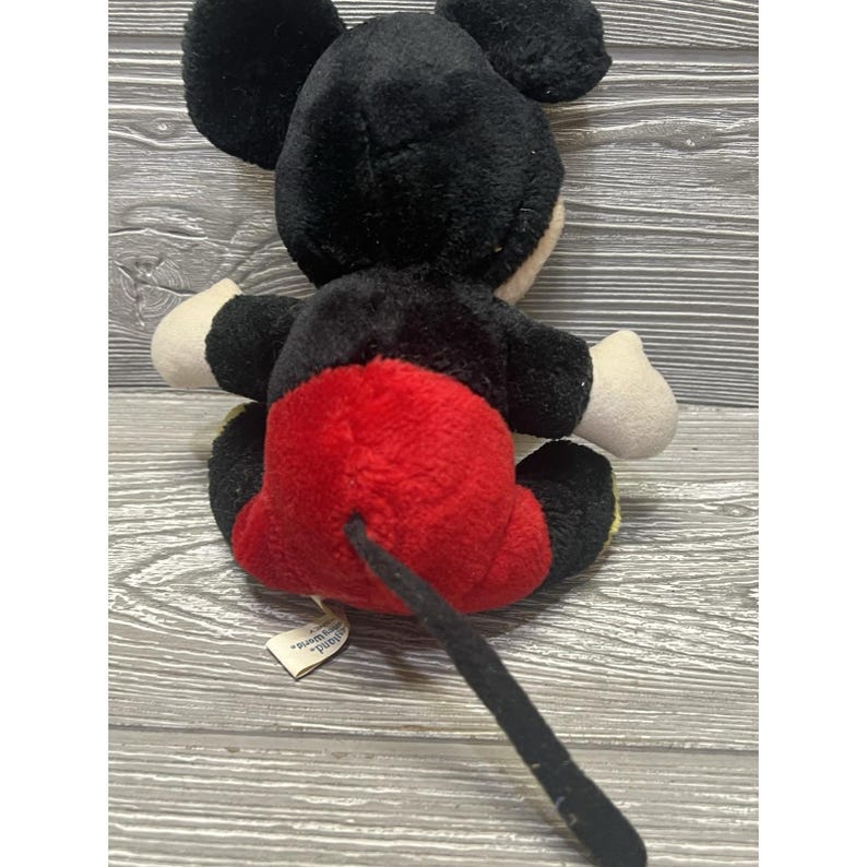 May include: A Mickey Mouse soft toy, seen from the rear. It has black fur, red shorts, white gloves, and a long black tail. The toy is positioned on a wooden surface.