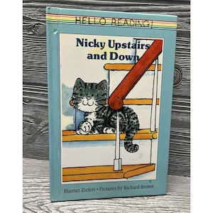 May include: A children's book titled "Nicky Upstairs and Down" with a cartoon illustration of a gray and white cat on a staircase. The book cover has a light blue border and the words "HELLO READING!" at the top.