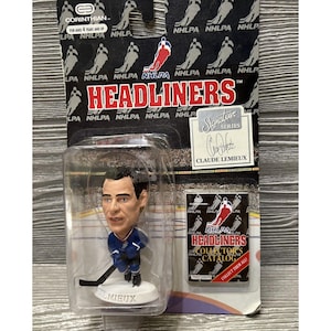 May include: A plastic figure of hockey player Claude Lemieux in a blue and white uniform, holding a hockey stick. The figure is part of the NHLPA Headliners Signature Series. The packaging features the NHLPA logo and the text "Headliners" in red.