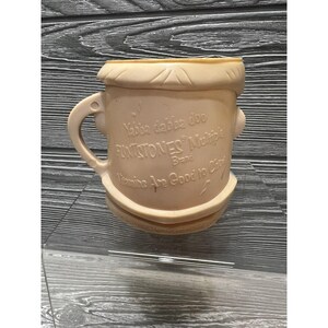 May include: A light pink plastic cup with a cartoon face and the words "Yabba dabba doo" and "Flintstones Multi-Vitamins Brand" printed on it. The cup has a handle and the text "Vitamins Are Good to Chew" is printed on the bottom.