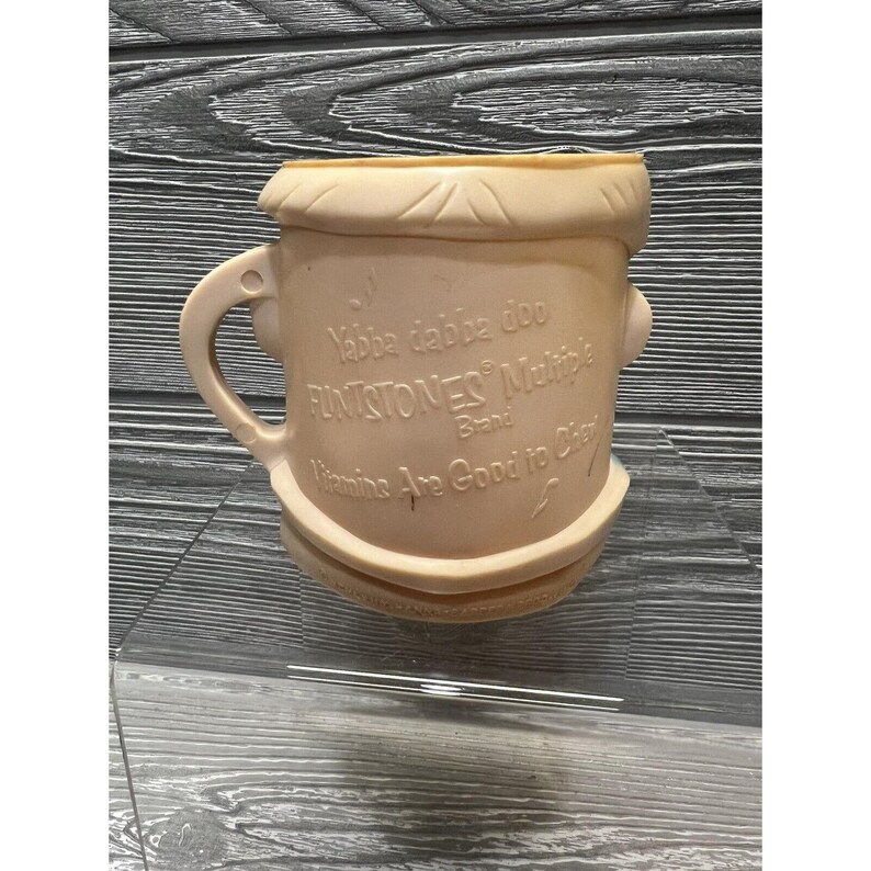 May include: A light pink plastic cup with a cartoon face and the words "Yabba dabba doo" and "Flintstones Multi-Vitamins Brand" printed on it. The cup has a handle and the text "Vitamins Are Good to Chew" is printed on the bottom.