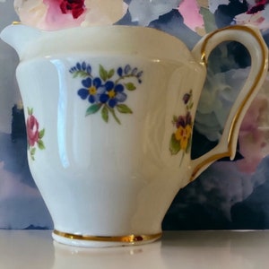 May include: A white porcelain creamer with a gold rim and floral pattern. The creamer has a small spout and a handle.