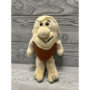 May include: A plush toy of a cartoon character with a brown loincloth and a big smile. The toy has a light brown body and a dark brown face with large black eyes.