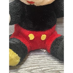 May include: Close-up of a plush Mickey Mouse toy. The toy has a black body, red shorts with yellow buttons, and yellow shoes. The face is white with a red mouth.