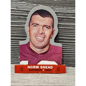May include: A vintage football card featuring Norm Snead, quarterback for the Eagles. The card has a cutout shape around the player's head and shoulders. The player is wearing a maroon jersey. The card's base has the player's name and team.
