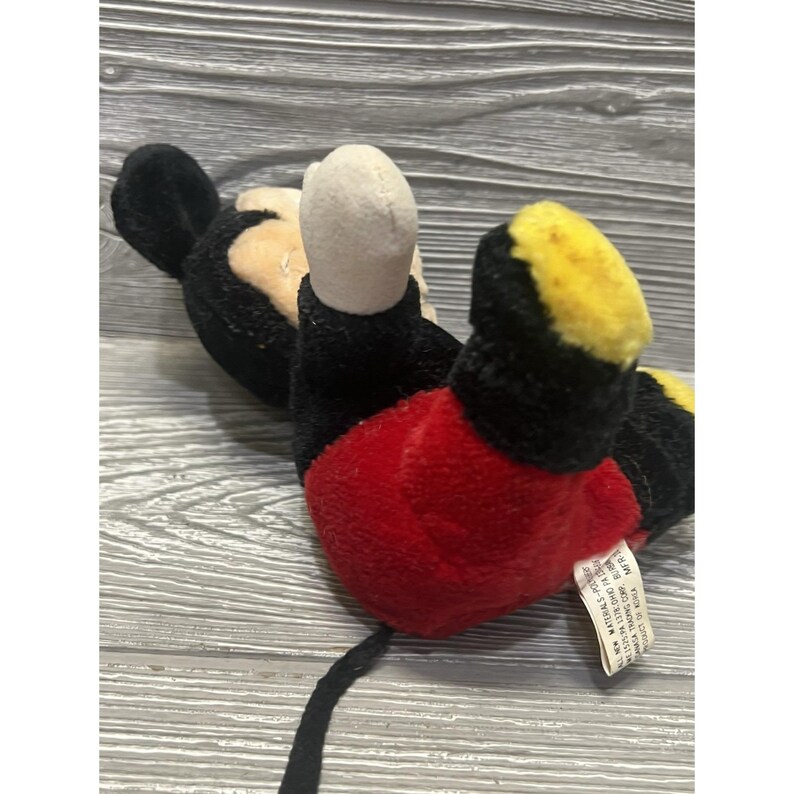 May include: A plush Mickey Mouse toy, with a black body, ears, and tail, red shorts, yellow shoes, and a beige face. The toy is lying on its back. A white tag is attached to the toy.