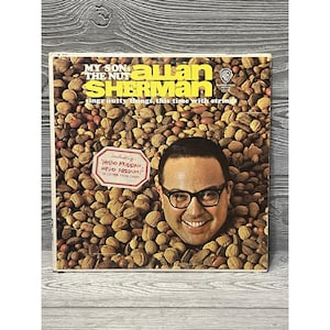 May include: Vintage vinyl record album cover featuring Allan Sherman's "My Son, The Nut." The cover art shows Sherman's face emerging from a pile of nuts. The album title and artist's name are prominently displayed, along with the Warner Bros. logo.