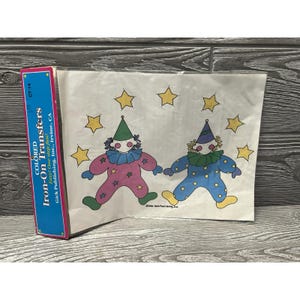 May include: A package of colored iron-on transfers featuring two cartoon clowns with star accents. The clowns are wearing colorful outfits and hats. The package is blue with white text that reads "COLORED Iron-On Transfers".