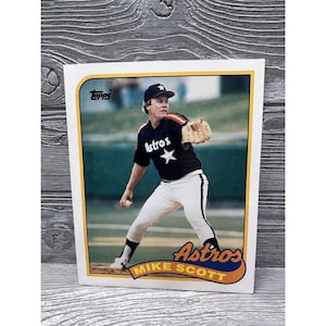 May include: A vintage Topps baseball card featuring Mike Scott, a pitcher for the Houston Astros. The card is white with a yellow border and features a photo of Scott in his Astros uniform, throwing a baseball. The card also includes the team logo and Scott's name.
