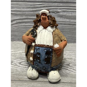 May include: A ceramic figurine of a portly man wearing a white cap, shirt, and shoes, with a brown jacket and blue pants. The figure is holding a wrench and a small white object.