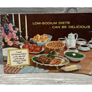 May include: A vintage advertisement for low-sodium diets featuring a table set with a floral centerpiece, a basket of rolls, a pie, a teapot, cups, a platter of meat, green beans, and a card reading A special Diet Guide and Cookbook.