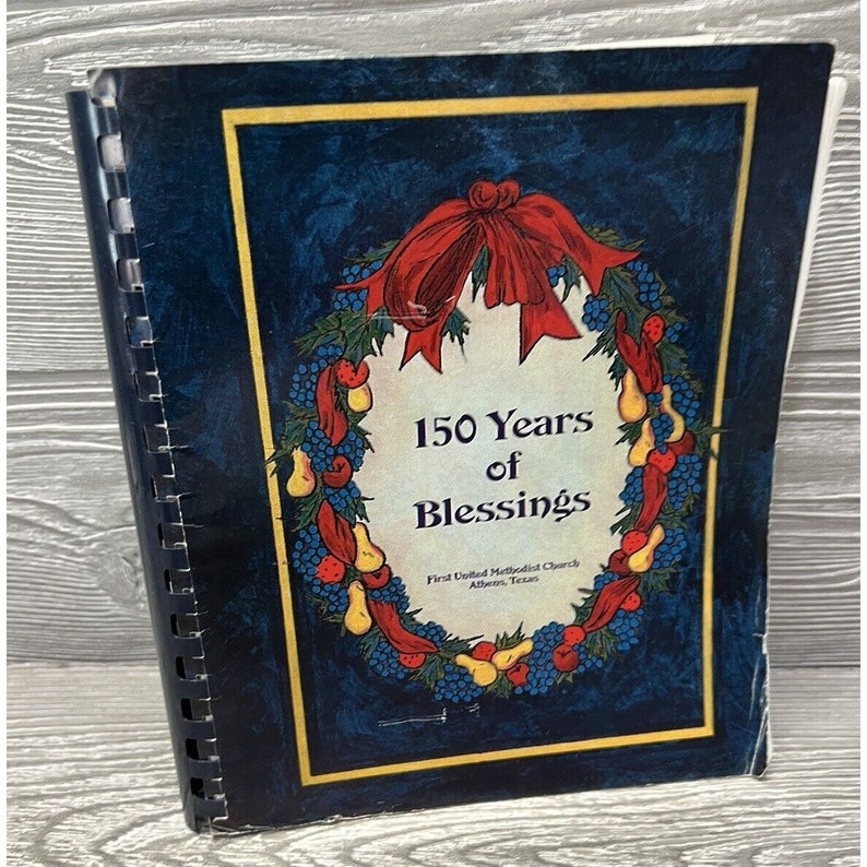 May include: A blue book with a gold border and a wreath of red berries, yellow pears, and blue grapes. The book has the title "150 Years of Blessings" and the text "First United Methodist Church Athens, Texas".