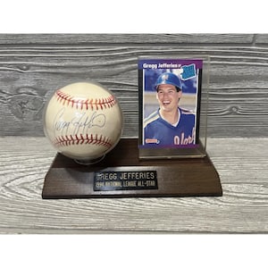 May include: A baseball signed by Gregg Jefferies, displayed with a baseball card in a clear case. The baseball and card are on a dark wooden base with a plaque that reads "Gregg Jefferies 1994 National League All-Star."