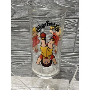 May include: A clear glass drinking glass with a Cabbage Patch Kids graphic. The graphic features a boy with yellow shorts and a red shirt, upside down, with two girls in red dresses on either side of him. The text "Cabbage Patch Kids" is printed in a black circle above the children.