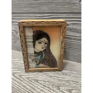 May include: A rectangular wooden frame with a carved scalloped edge containing a miniature painting of a person with dark hair, wearing a purple head covering and blue garment, standing beside a white pillar against a soft orange background.