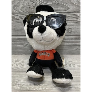 May include: A black and white plush panda wearing a black baseball cap and sunglasses. The panda is wearing an orange and black shirt with the word "Giants" printed on it.