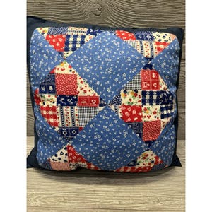 May include: A square pillow with a patchwork design. The pillow features a blue background with a floral pattern, and a central design of red and blue squares. The pillow is approximately 16 inches square.