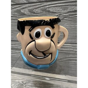 May include: A vintage plastic mug shaped like Fred Flintstone's head. The mug is light brown with a blue collar and black hair. Fred has a big smile and large eyes.