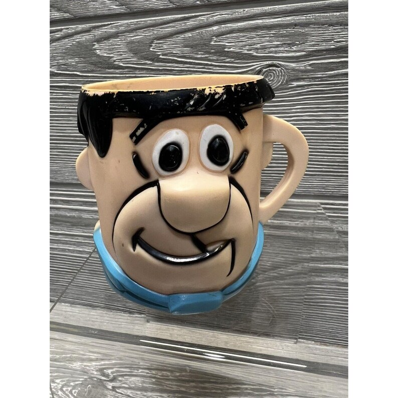 May include: A vintage plastic mug shaped like Fred Flintstone's head. The mug is light brown with a blue collar and black hair. Fred has a big smile and large eyes.
