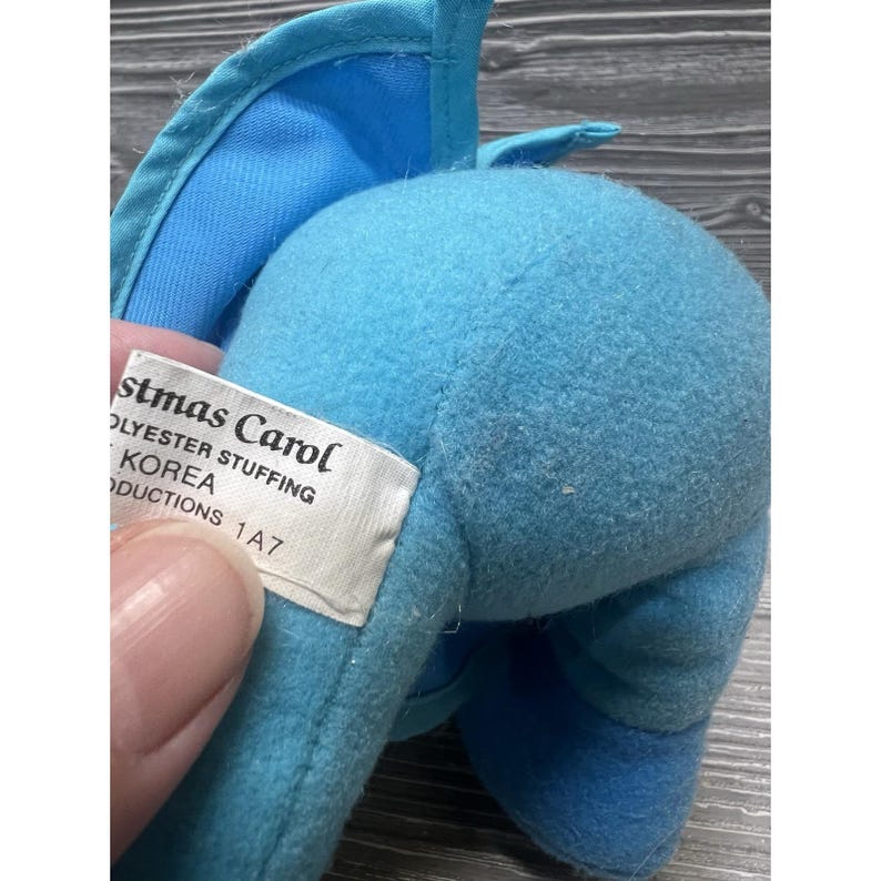 Pode incluir: Um close-up de um brinquedo de pel&uacute;cia azul com uma etiqueta que diz "Christmas Carol". O brinquedo &eacute; feito de material macio e difuso e tem uma asa azul clara. A etiqueta tamb&eacute;m indica "Enchimento de poli&eacute;ster" e "Korea Productions 1A7."