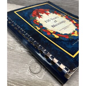 May include: A blue book with a gold border and a colorful wreath on the cover. The wreath is made of fruit, berries, and leaves. The book's title is "150 Years of Blessings".