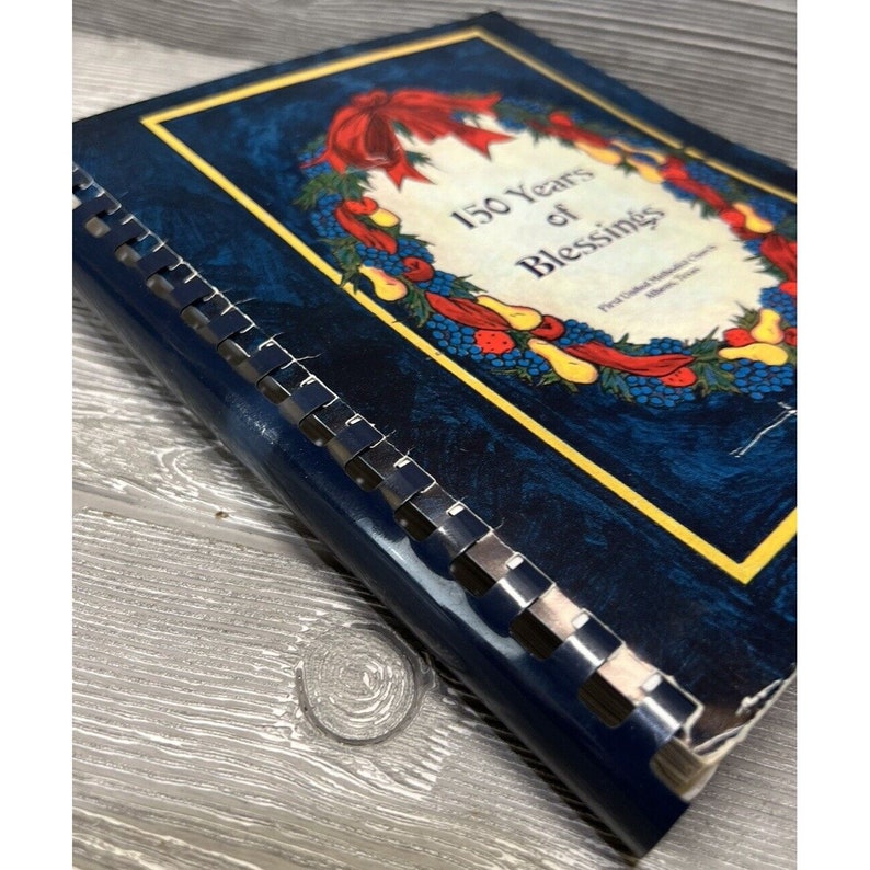 May include: A blue book with a gold border and a colorful wreath on the cover. The wreath is made of fruit, berries, and leaves. The book's title is "150 Years of Blessings".