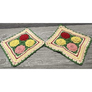 May include: Two square crochet potholders featuring a floral motif. Each piece displays four raised, three-dimensional flowers in red, yellow, and pink, surrounded by a multi-colored border of cream, yellow, red, and green yarn stitches.
