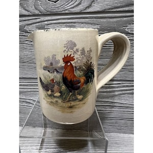 May include: A cream-colored ceramic pitcher with a rooster and chicks design. The rooster is red, black, and white, and the chicks are yellow. The pitcher has a handle and a rustic, farmhouse style.