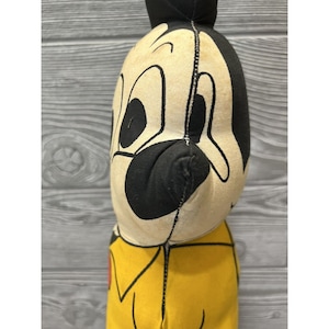 May include: Vintage plush toy of a cartoon character with a black nose, large eyes, and a yellow shirt. The toy is made of fabric and has a worn appearance.