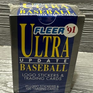 May include: A sealed box of Fleer Ultra Update Baseball trading cards from 1991. The box is primarily navy blue with yellow and white text. It includes logo stickers and trading cards. The box contains 20 logo stickers and 120 trading cards.