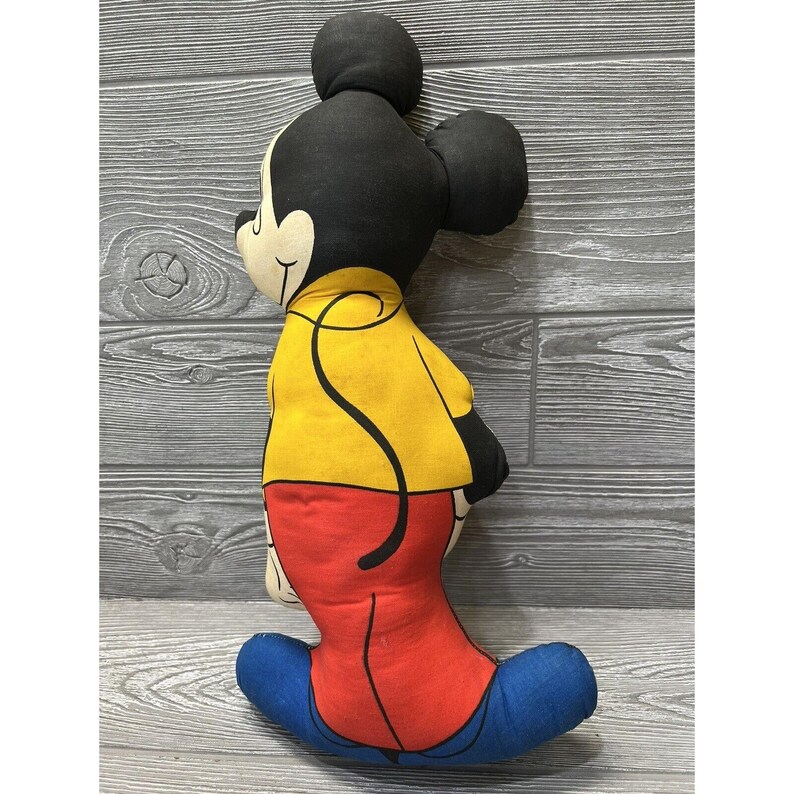 May include: A vintage Mickey Mouse plush toy with a yellow shirt, red pants, and blue shoes. The toy is made of fabric and has a classic cartoon design.