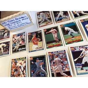 May include: A collection of 1991 Topps baseball cards, including cards of Don Mattingly, Chipper Jones, and Ryne Sandberg. The cards are in a box labeled "1991 Topps Baseball Near Complete Hand Set". Some cards are labeled "Record Breaker" and "All Star".