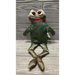 May include: A green satin frog plush toy with pink eye makeup and red lips. The frog has long legs and arms, and a tag is attached to its body. The plush is sitting on a gray wooden surface.