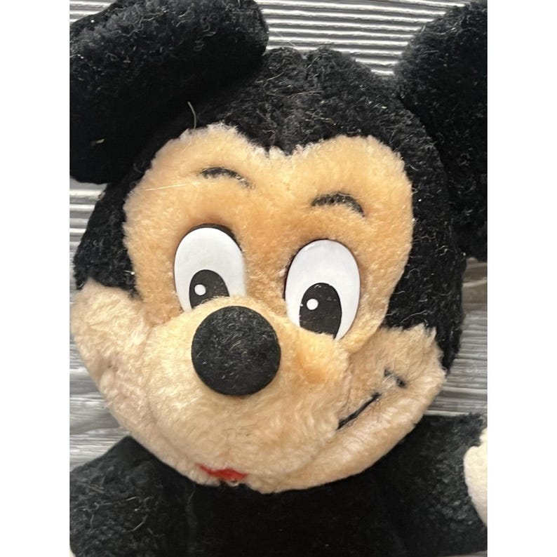 May include: Close-up of a vintage Mickey Mouse plush toy. The plush has a black body and ears, with a smiling face featuring a tan muzzle, large white eyes, and a black nose. The toy is made of soft, fuzzy material.