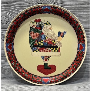 May include: A round, cream-colored metal serving tray with a red and green border of hearts. The center features a whimsical illustration of Santa Claus holding a bird and a bag of gifts.