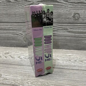 May include: Two VHS tapes with a purple and green design. The tapes are labeled "Doo Wop 5" and feature a group of singers on the cover. The text on the tapes reads "Volume One" and "Volume Two".