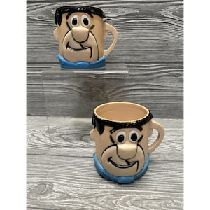 May include: Two vintage plastic cups featuring the cartoon character Fred Flintstone. The cups are beige with black hair and a blue shirt.