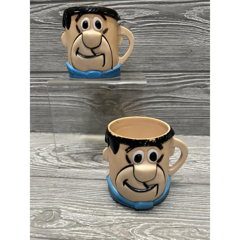 May include: Two vintage plastic cups featuring the cartoon character Fred Flintstone. The cups are beige with black hair and a blue shirt.