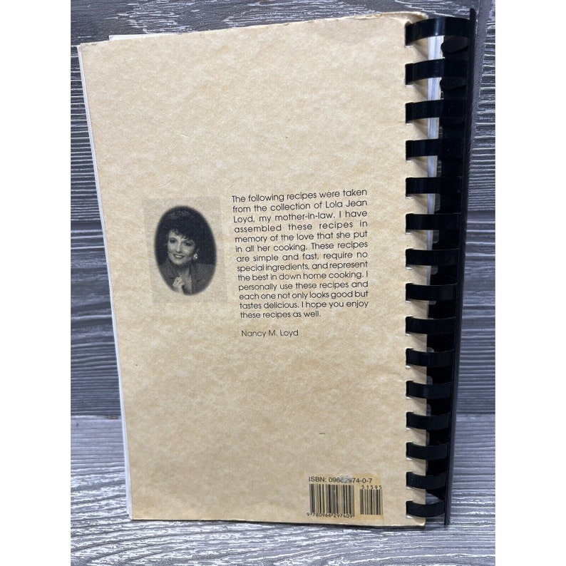 Puede incluir: Un libro de cocina vintage con una cubierta de color beige y una encuadernaci&oacute;n en espiral negra. La cubierta presenta una foto de una mujer y un texto que dice "The following recipes were taken from the collection of Lola Jean Loyd, my mother-in-law. I have assembled these recipes in memory of the love that she put in all her cooking. These recipes are simple and fast, require no special ingredients, and represent the best in down home cooking. I personally use these recipes and each one not only looks good but tastes delicious. I hope you enjoy these recipes as well. Nancy M. Loyd" y "ISBN: 09602974-0-7".