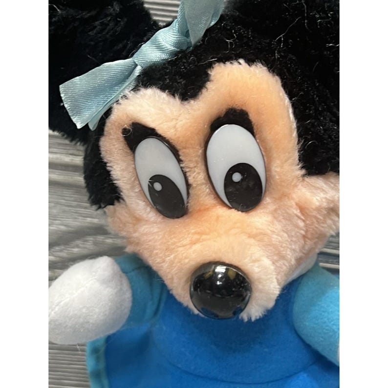 Pode incluir: Close-up de um brinquedo de pel&uacute;cia com um personagem de desenho animado com pelo preto, olhos brancos grandes e um nariz preto. O personagem tem um la&ccedil;o azul claro e est&aacute; usando um vestido azul. O rosto do brinquedo &eacute; cor de p&ecirc;ssego.