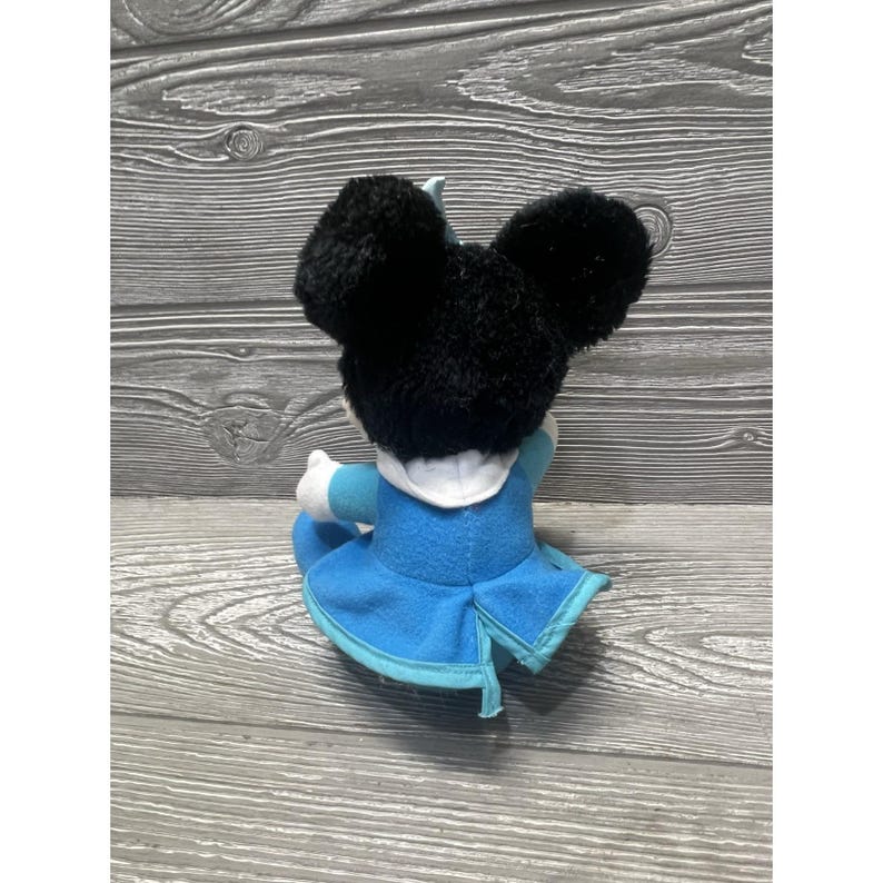 Pode incluir: Um brinquedo de pel&uacute;cia de um personagem com grandes orelhas pretas, vestindo um vestido azul claro com gola branca. O brinquedo est&aacute; sentado em uma superf&iacute;cie de madeira.