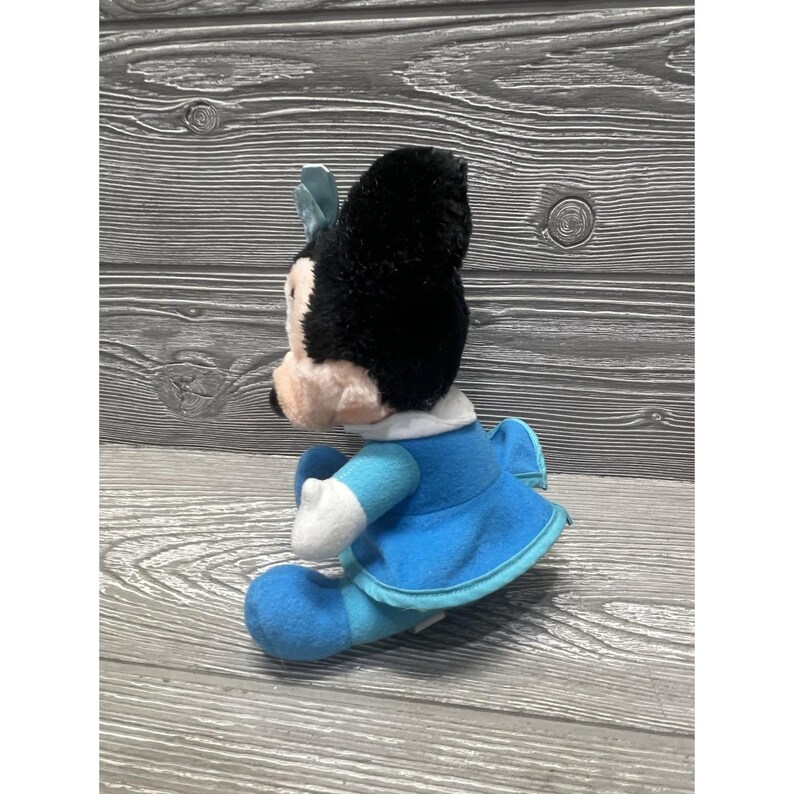 Pode incluir: Uma boneca de pel&uacute;cia da Minnie Mouse, com uma cabe&ccedil;a preta e um la&ccedil;o azul, uma gola branca e um vestido azul. A boneca tem luvas brancas e sapatos azuis. O brinquedo &eacute; feito de tecido macio e &eacute; projetado para crian&ccedil;as.