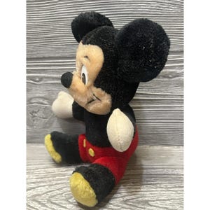 May include: A plush Mickey Mouse toy, with black fur, a tan face, and red shorts. The classic character has large black ears, white gloves, and yellow shoes. The vintage toy is sitting on a wooden surface.