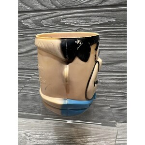 May include: A vintage plastic character head cup with a black and white face and a blue collar. The cup is made of a hard plastic and has a smooth surface.