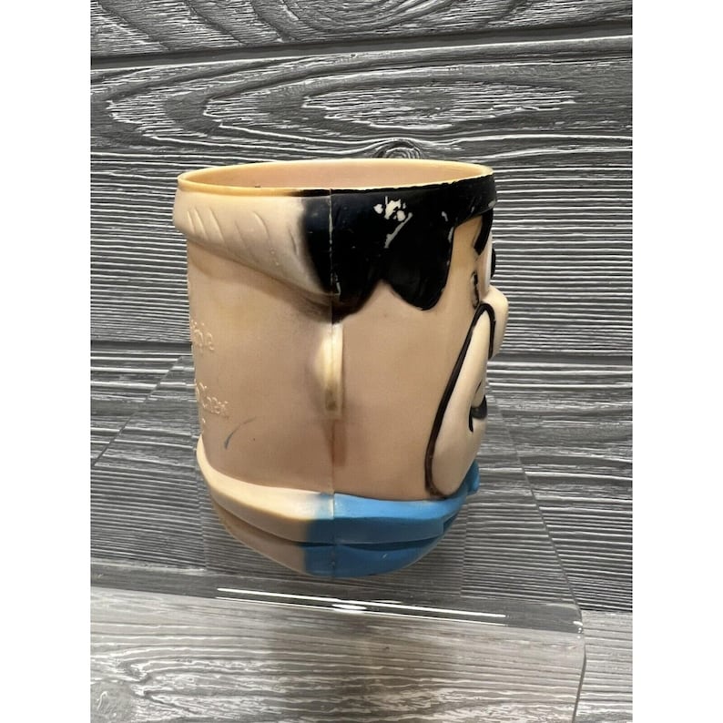 May include: A vintage plastic character head cup with a black and white face and a blue collar. The cup is made of a hard plastic and has a smooth surface.