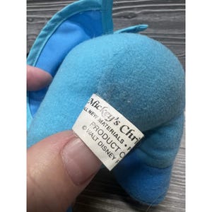 Pode incluir: Um brinquedo de pel&uacute;cia azul claro com uma etiqueta que diz "Mickey's Christmas". O brinquedo tem um corpo arredondado e uma orelha pontuda. A etiqueta tamb&eacute;m inclui o texto "ALL NEW MATERIALS" e "PRODUCT OF WALT DISNEY".