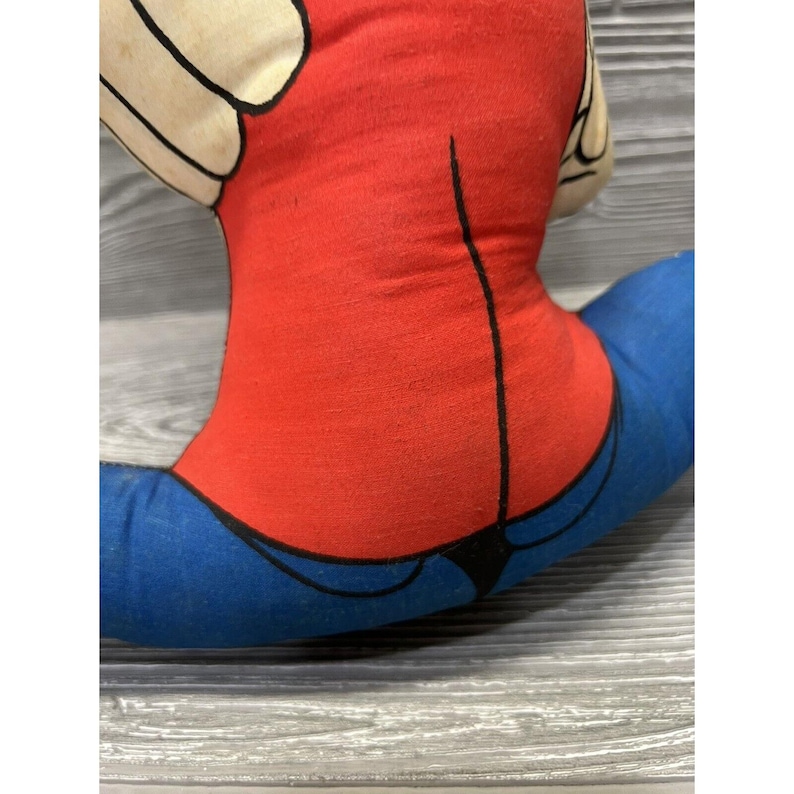May include: A red and blue plush toy in the shape of a person sitting with their legs crossed. The toy has a white face and black eyes.
