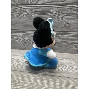 Pode incluir: Uma boneca de pel&uacute;cia da Minnie Mouse, com pelo preto, um la&ccedil;o azul claro e um vestido azul com detalhes brancos. A boneca est&aacute; sentada e tem uma gola branca. O fundo &eacute; uma superf&iacute;cie de madeira.