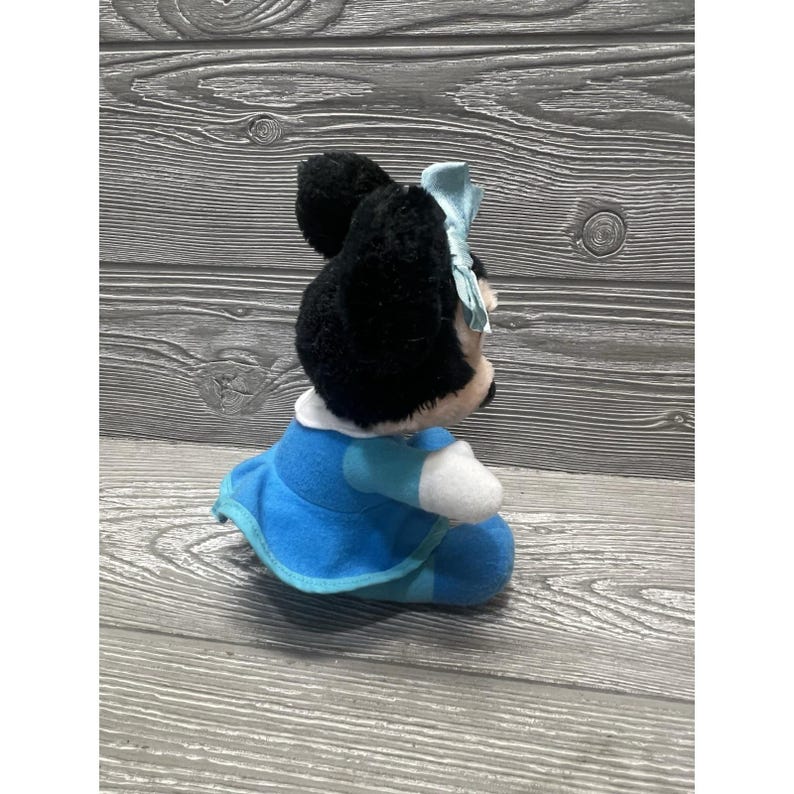 Pode incluir: Uma boneca de pel&uacute;cia da Minnie Mouse, com pelo preto, um la&ccedil;o azul claro e um vestido azul com detalhes brancos. A boneca est&aacute; sentada e tem uma gola branca. O fundo &eacute; uma superf&iacute;cie de madeira.